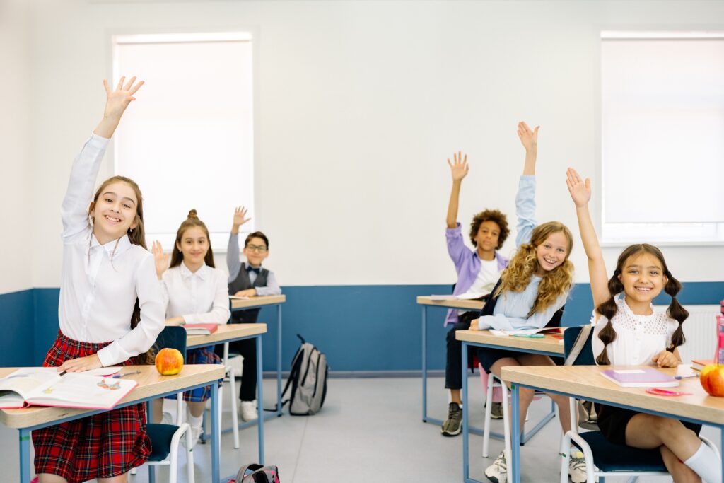 Group of diverse children eagerly raising hands in a bright classroom setting.
